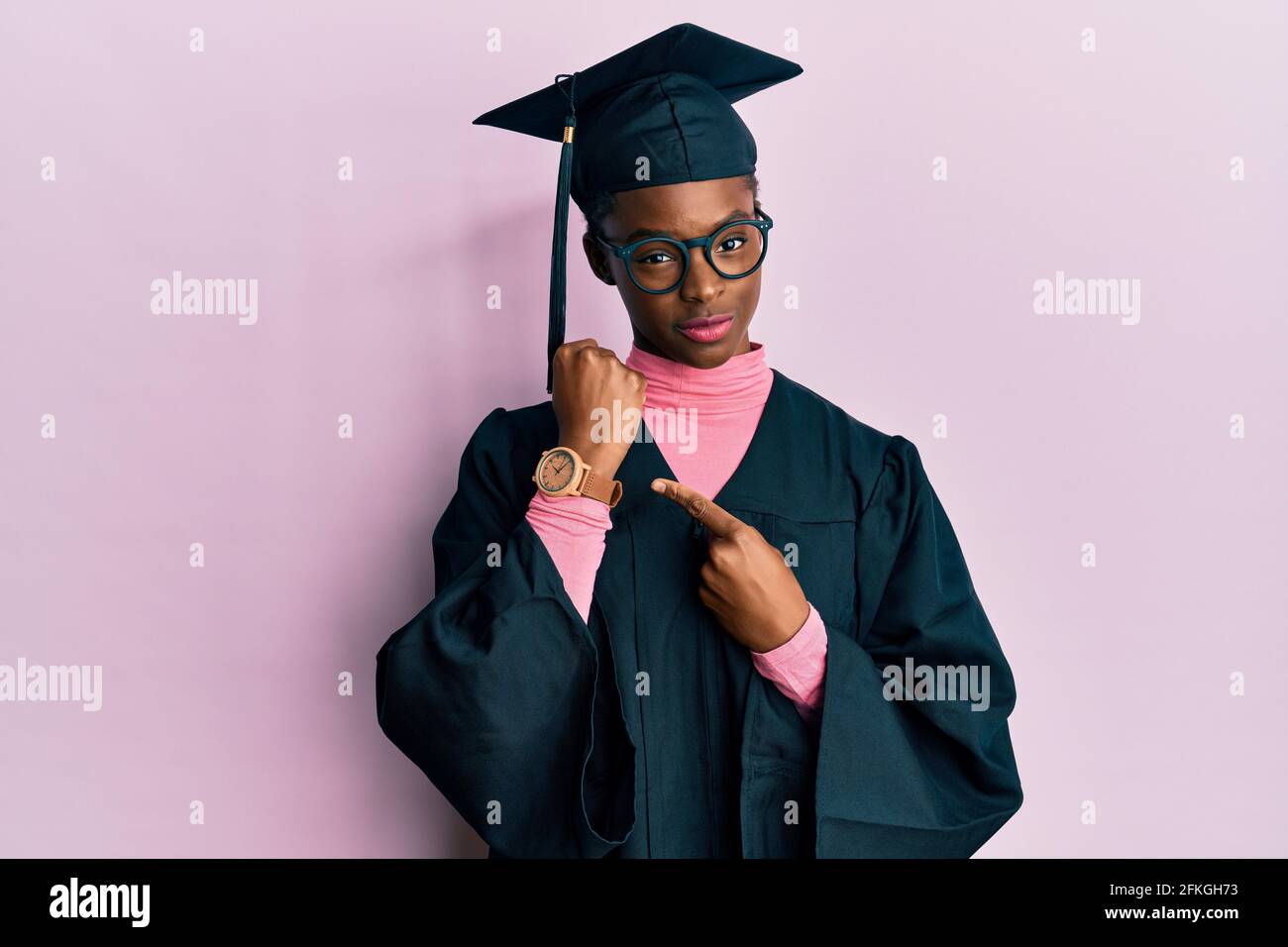 Young african american girl wearing graduation cap and ceremony robe in hurry pointing to watch