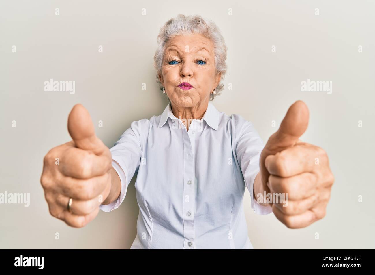 Senior grey-haired woman doing thumbs up positive gesture puffing cheeks with funny face. mouth inflated with air, catching air. Stock Photo