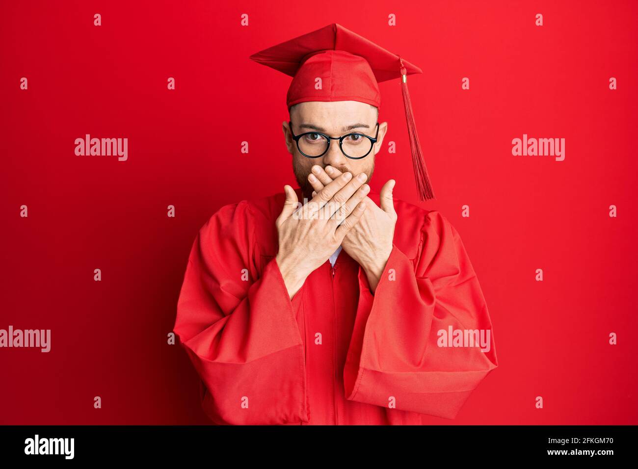 Young redhead man wearing red graduation cap and ceremony robe shocked ...