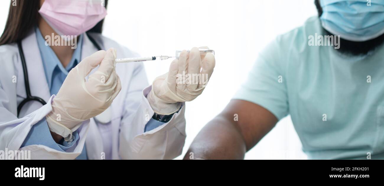 Asian doctor user syringe needle for injection vaccinate,  African American is being covid vaccinated for prevention coronavirus and flu. Concept of i Stock Photo