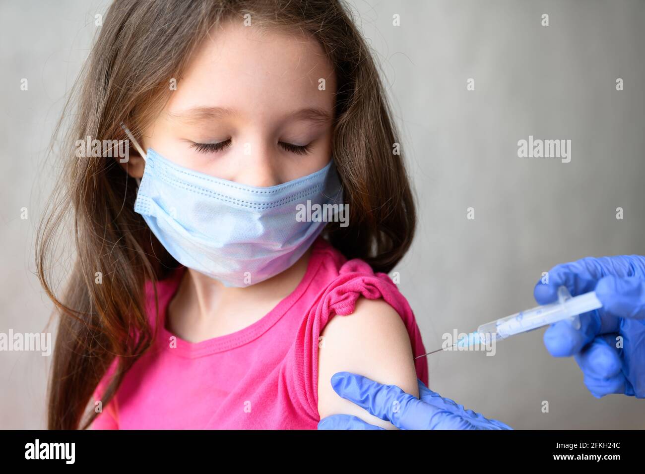 Vaccination of kid from COVID-19 or flu, cute little girl in mask closes eyes during injection. Doctor holds syringe for jab to child. Concept of coro Stock Photo