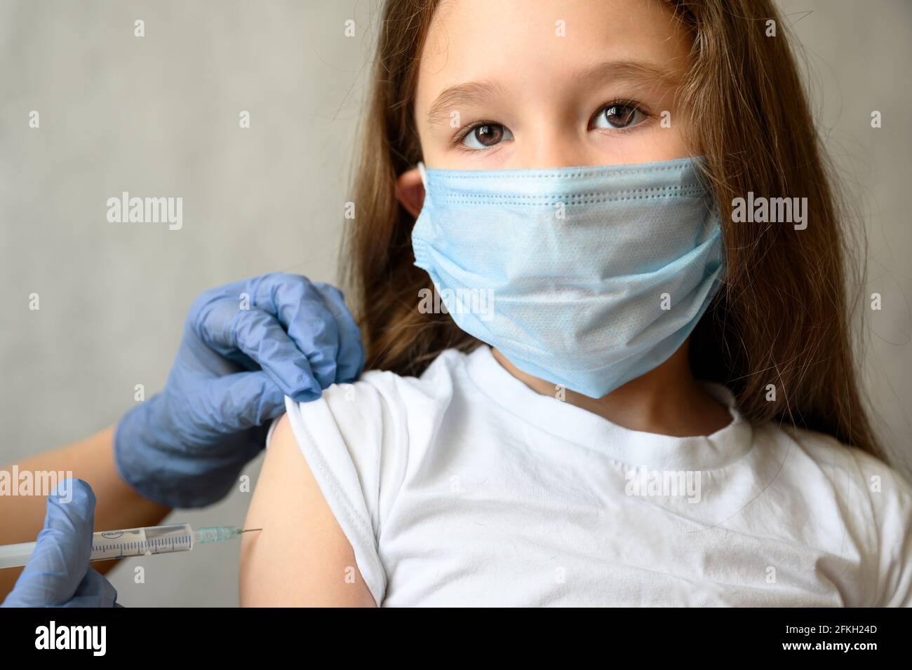 Vaccination of kid from COVID-19 or flu, pretty little girl looks at camera during coronavirus vaccine injection. Cute child face in mask and syringe. Stock Photo