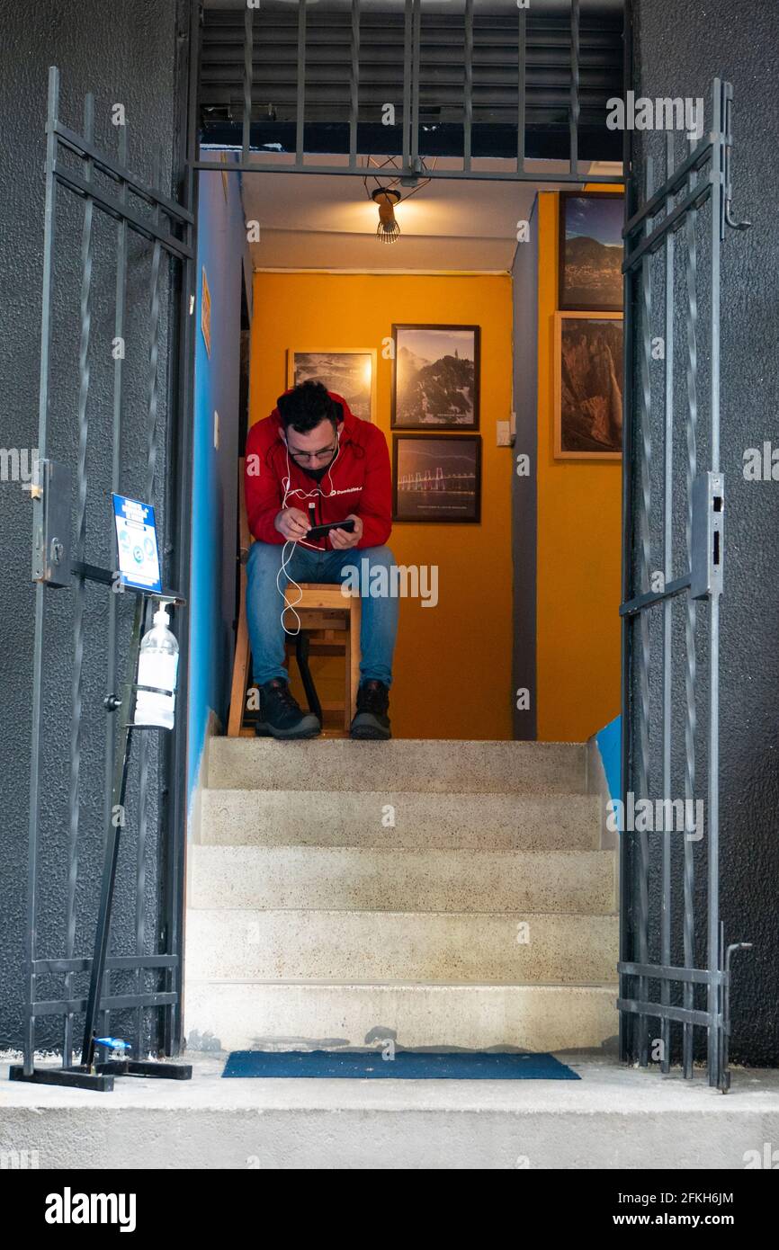 Medellin, Colombia - March 28 2021: Young Man Wearing a Black Mask is Sitting at the Entrance Looking at his Cell Phone Stock Photo