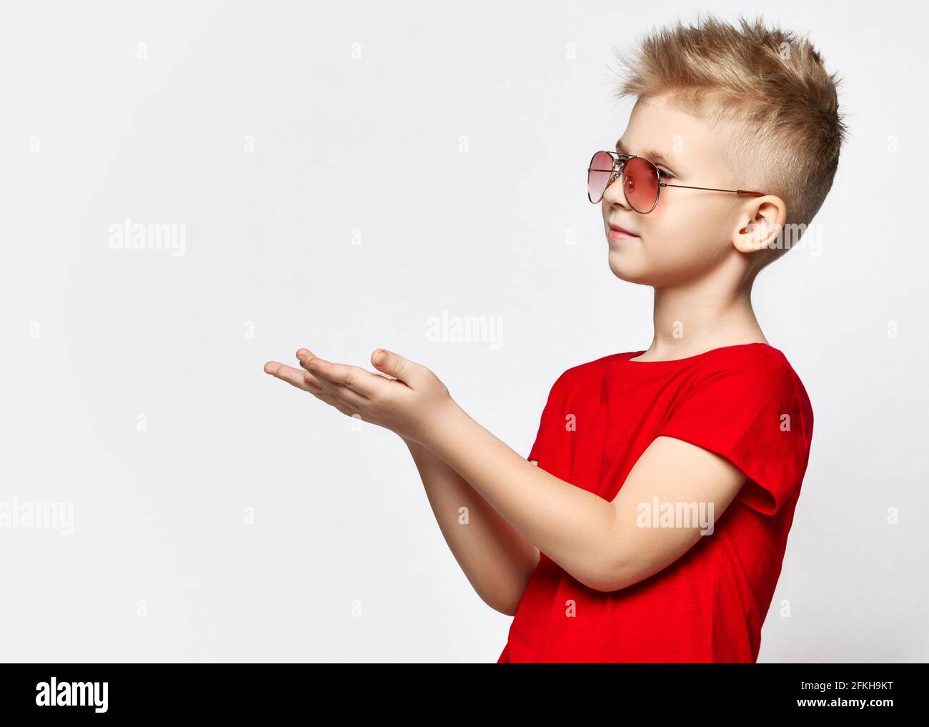 Portrait of blonde kid boy in red t-shirt and sunglasses standing holding hands up with open palms, holding blank space Stock Photo