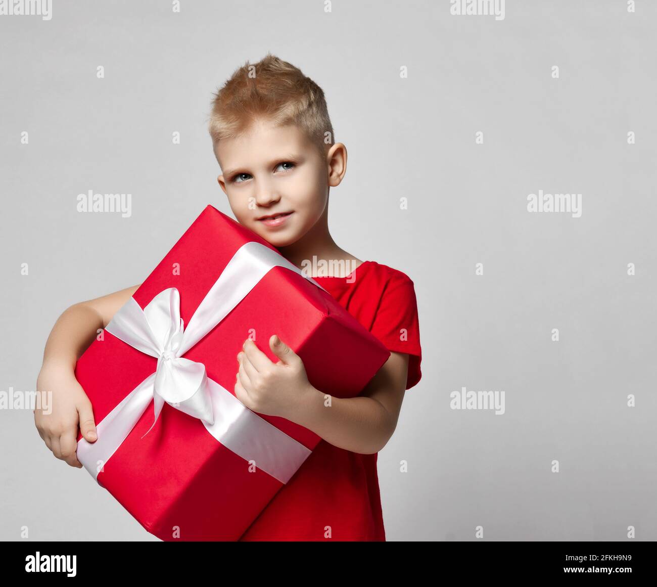 Portrait of cheerful kid boy in red t-shirt holding big present box with ribbon in hands, carrying it Stock Photo