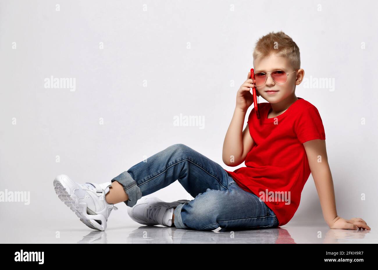 Cool blonde kid boy in red t-shirt, blue jeans, white sneakers and sunglasses sits on floor talking by cellphone Stock Photo