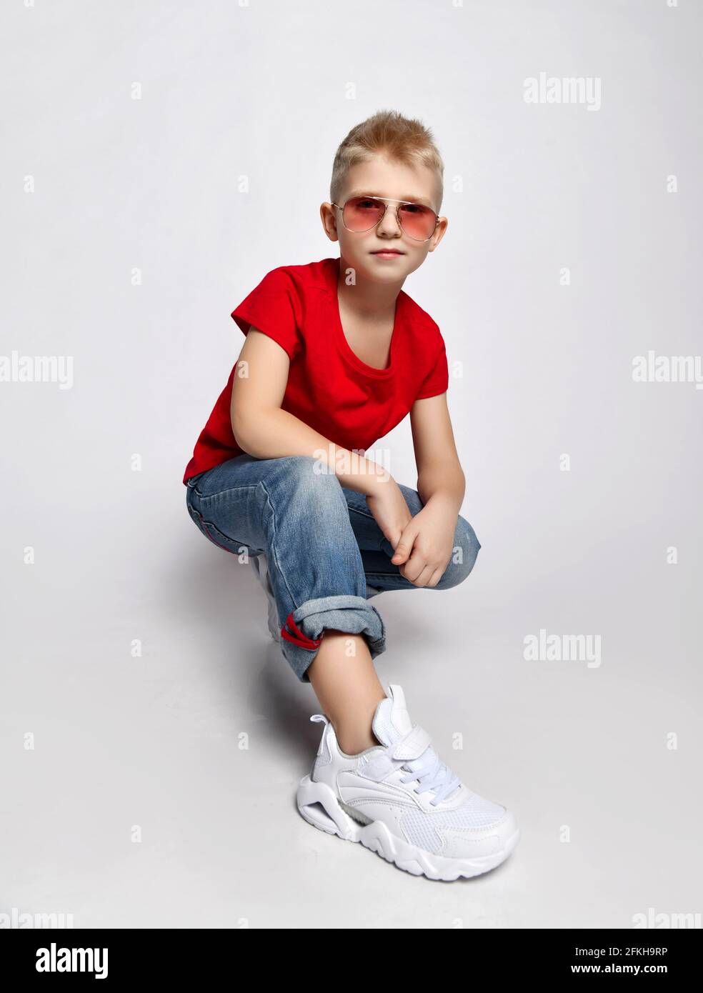 Self-confident blond kid boy in red t-shirt, blue jeans, sneakers and sunglasses sits squatted looking at camera Stock Photo