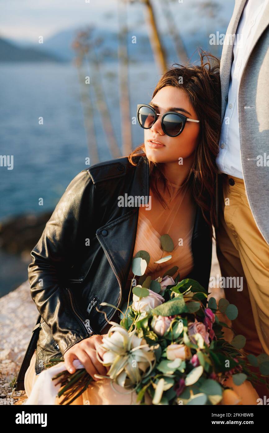 Bride in a leather jacket and sunglasses with a bouquet of flowers is ...