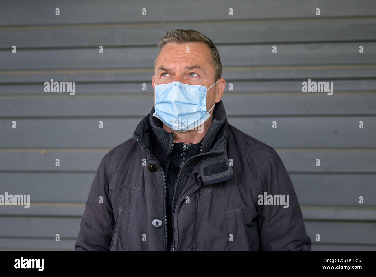 Thoughtful man wearing a protective face mask glancing to the side with a thoughtful expression as he poses outdoors against a grey wall during the Co Stock Photo