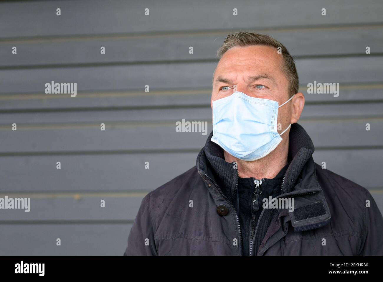 Thoughtful man wearing a protective face mask glancing to the side with a thoughtful expression as he poses outdoors against a grey wall during the Co Stock Photo