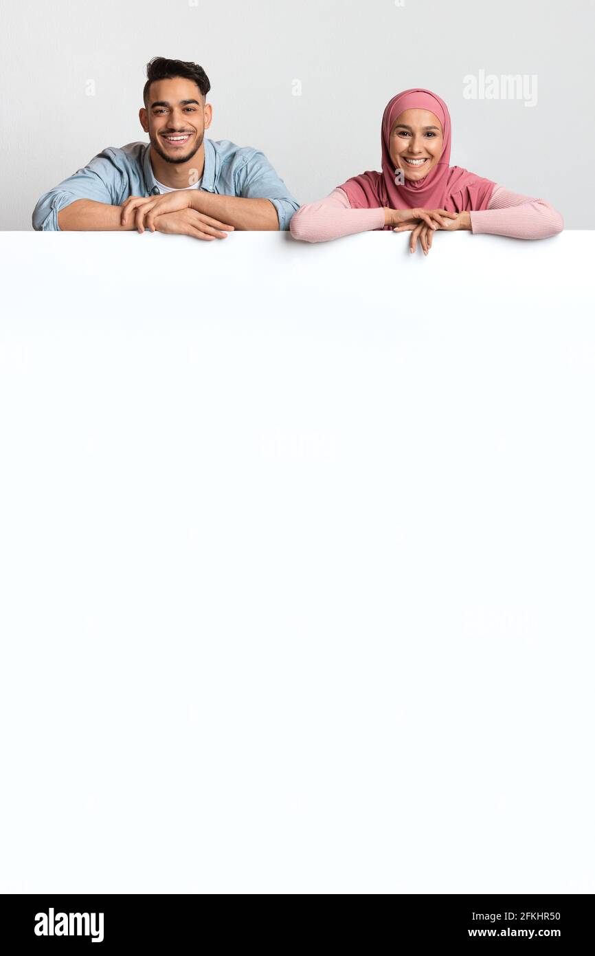 Nice Offer. Cheerful Muslim Spouses Leaning At Blank White Advertisement Board Stock Photo