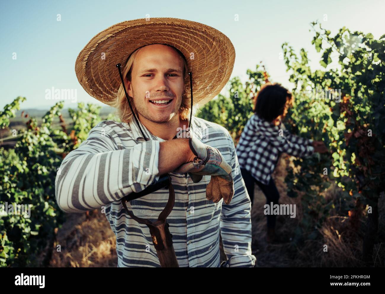 Caucasian blonde male farmer smiling in vineyards wearing straw hat leaning on pitch fork Stock Photo