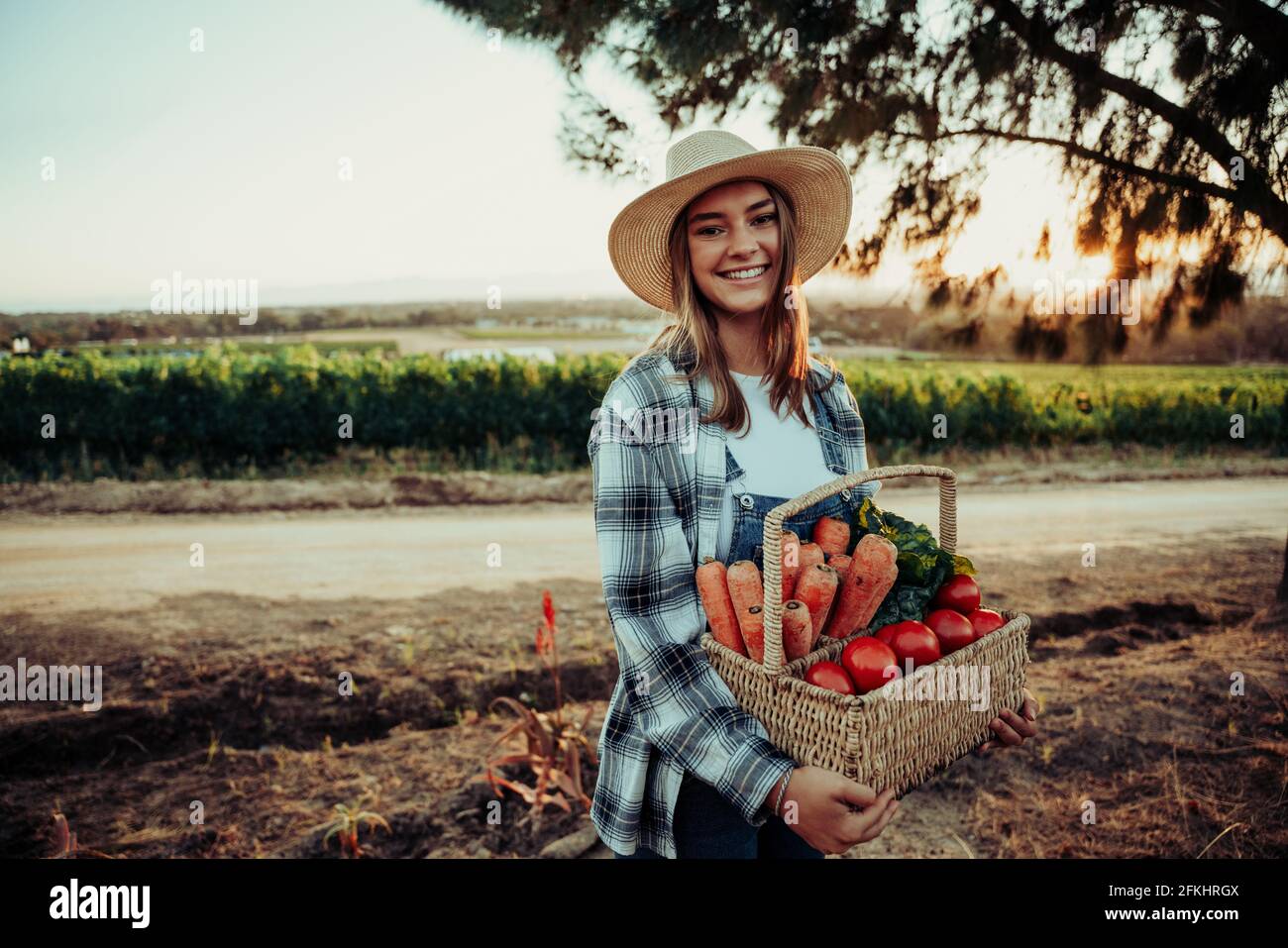 Smiling caucasian female farmer standing in fields holding basket of fresh vegetables  Stock Photo