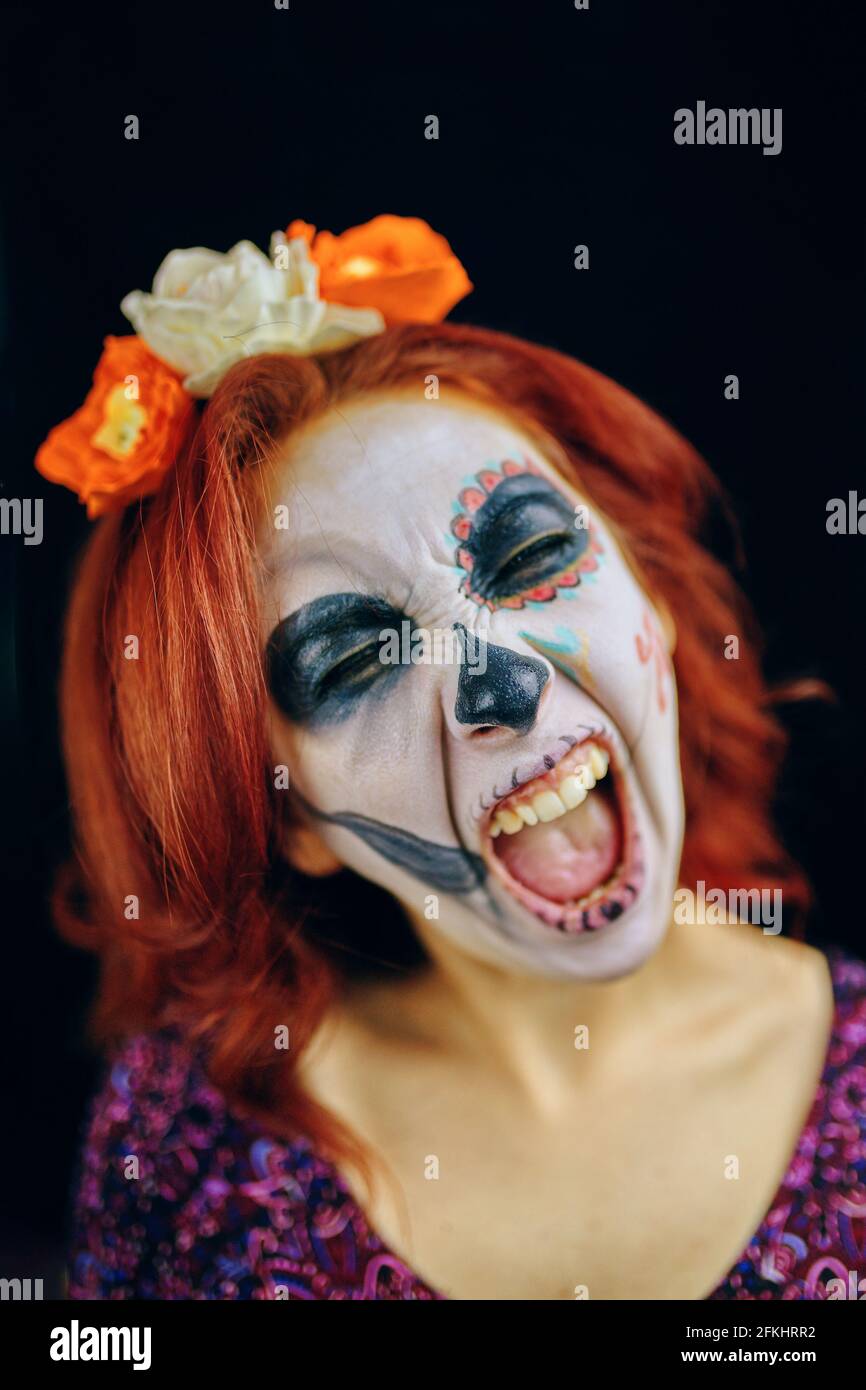 Young woman in day of the dead mask skull face art makeup and red hair, screaming on dark background close up Stock Photo