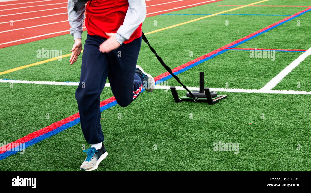 Front view of a high school runner pulling a sled with weight on it on a green turf field next to a red track at practice. Stock Photo