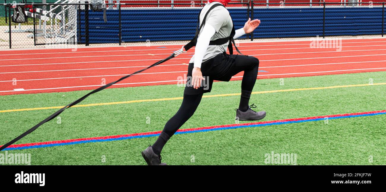 A high school teenage boy is running on a turf field with the bleachers in the background pulling a sled with weight during track practice. Stock Photo
