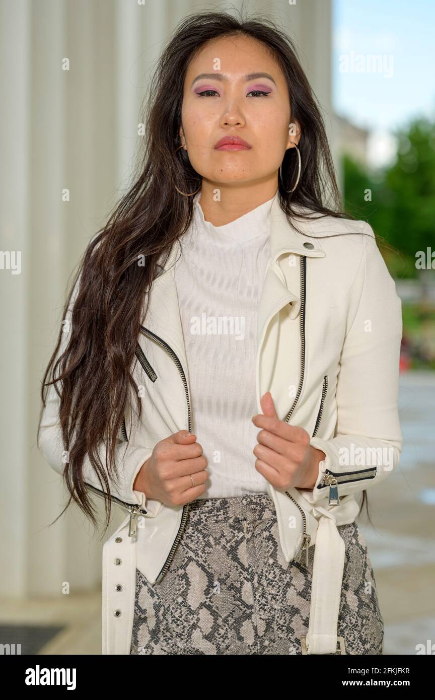 A young pretty Mongolian woman with a white leather jacket and patterned well-fitting pants in an urban park. Stock Photo