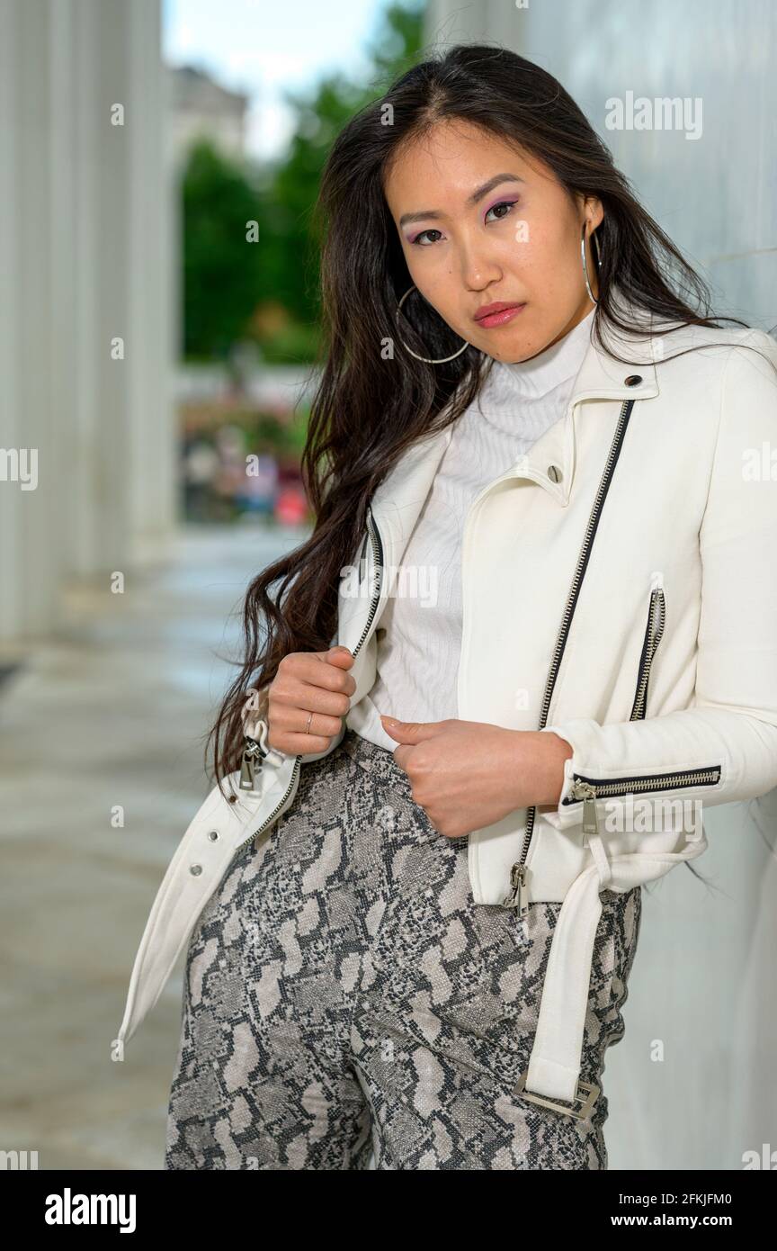 A young pretty Mongolian woman with a white leather jacket and patterned well-fitting pants in an urban park. Stock Photo