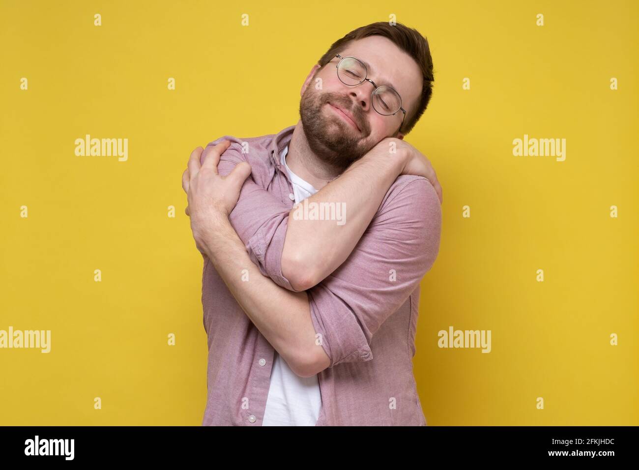 Cute, funny bearded man in round glasses hugs himself, he dreams of love or suffers from narcissism, closes eyes and smiles sweetly.  Stock Photo