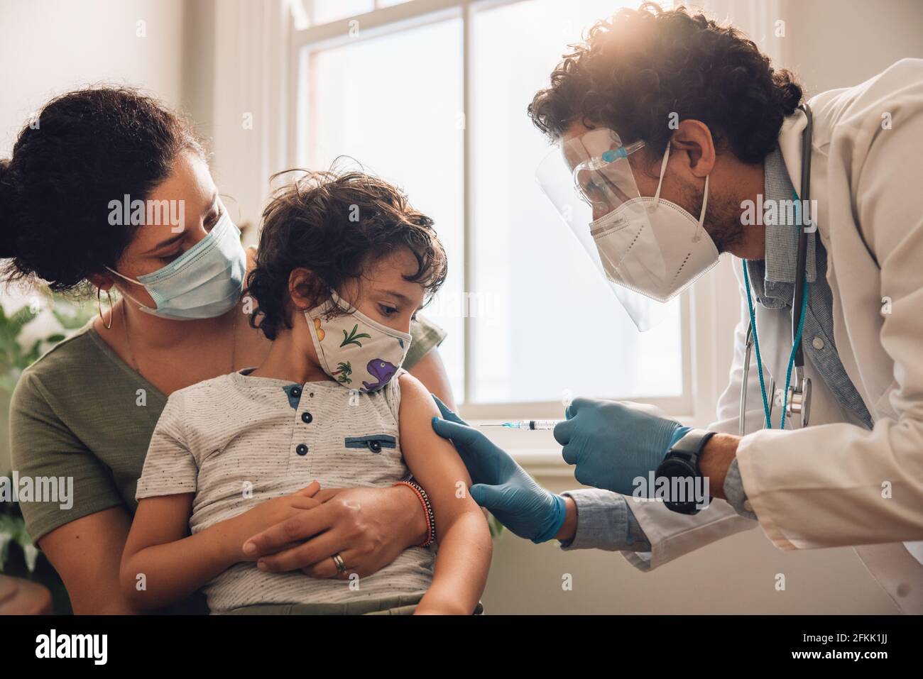 Frontline worker giving covid-19 vaccine injection to small boy sitting with her mother at home. Kid getting vaccinated at home during pandemic. Stock Photo