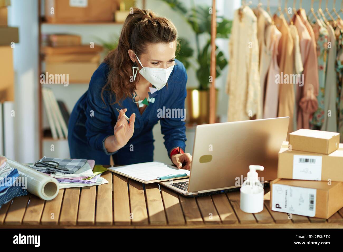 trendy 40 years old small business owner woman with ffp2 mask, parcel and antiseptic having video conference on laptop in the office. Stock Photo