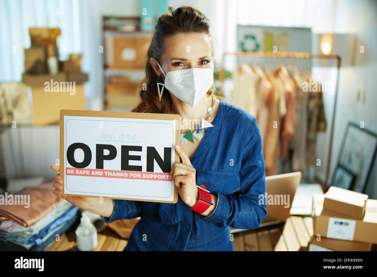 trendy middle aged small business owner woman with ffp2 mask and open after covid sign in the office. Stock Photo