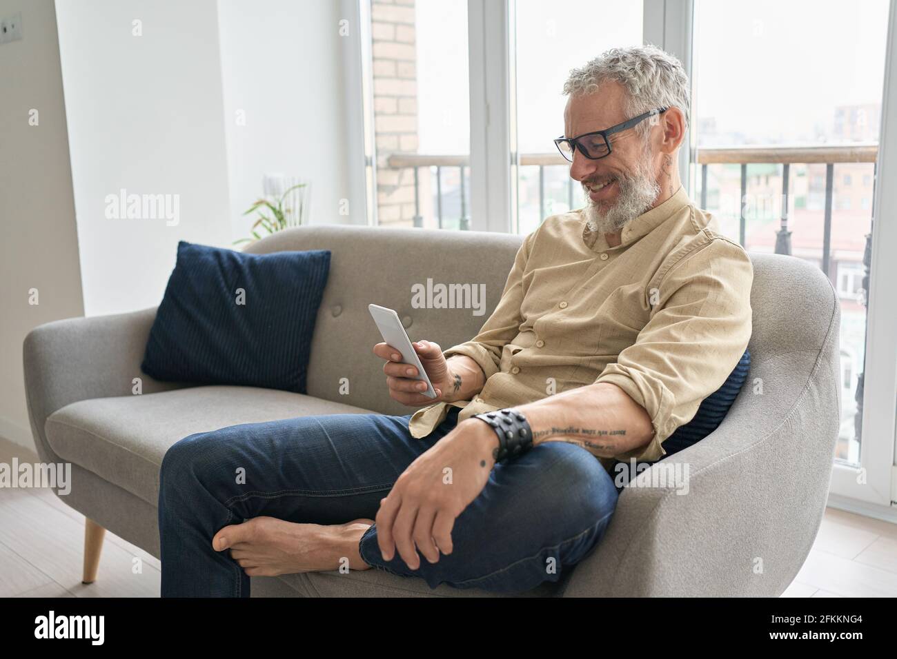 Happy older middle aged man using phone relaxing sitting on couch at home. Stock Photo