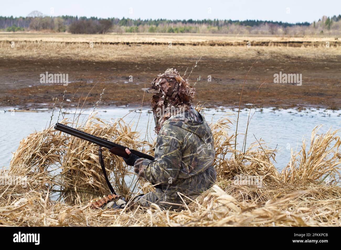 duck hunting in camouflage with a shotgun sitting next to the lake during waterfowl hunting Stock Photo
