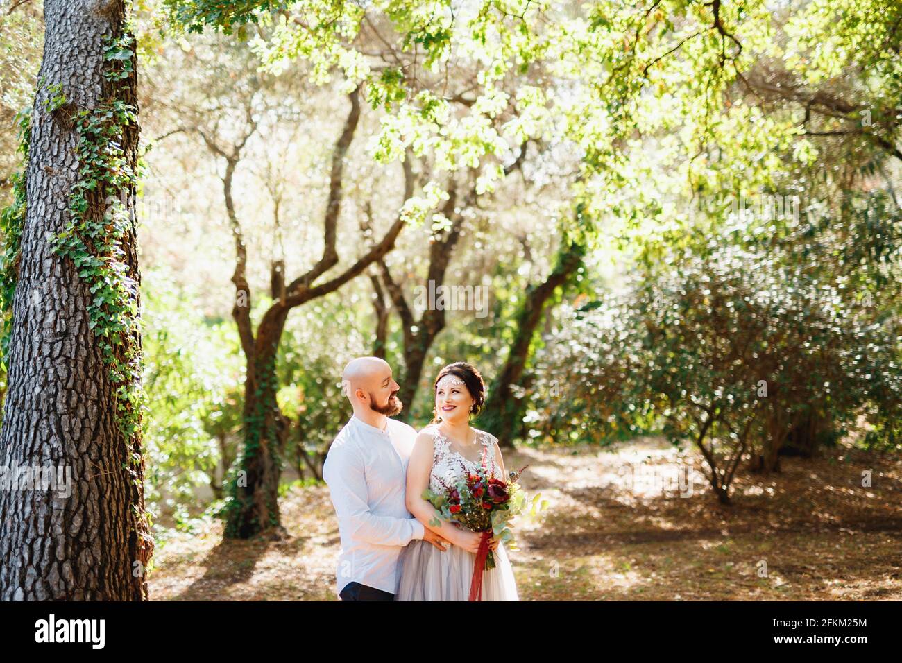 The bride and groom with a bouquet stand hugging among the trees in an ...