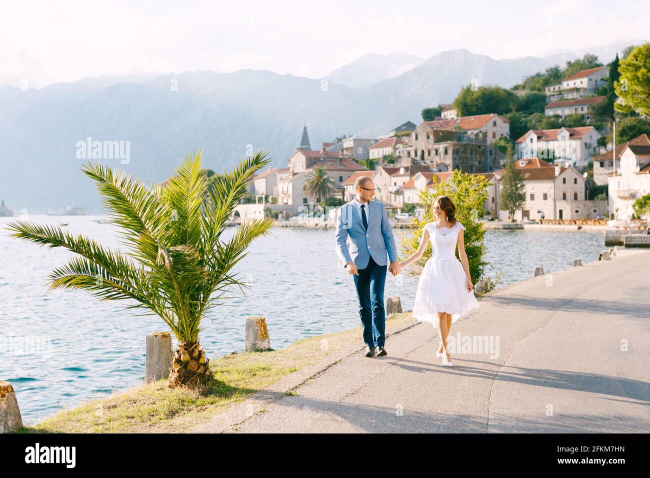 Smiling bride and groom walk along the road holding hands against the background of buildings of the city of Perast in Montenegro Stock Photo