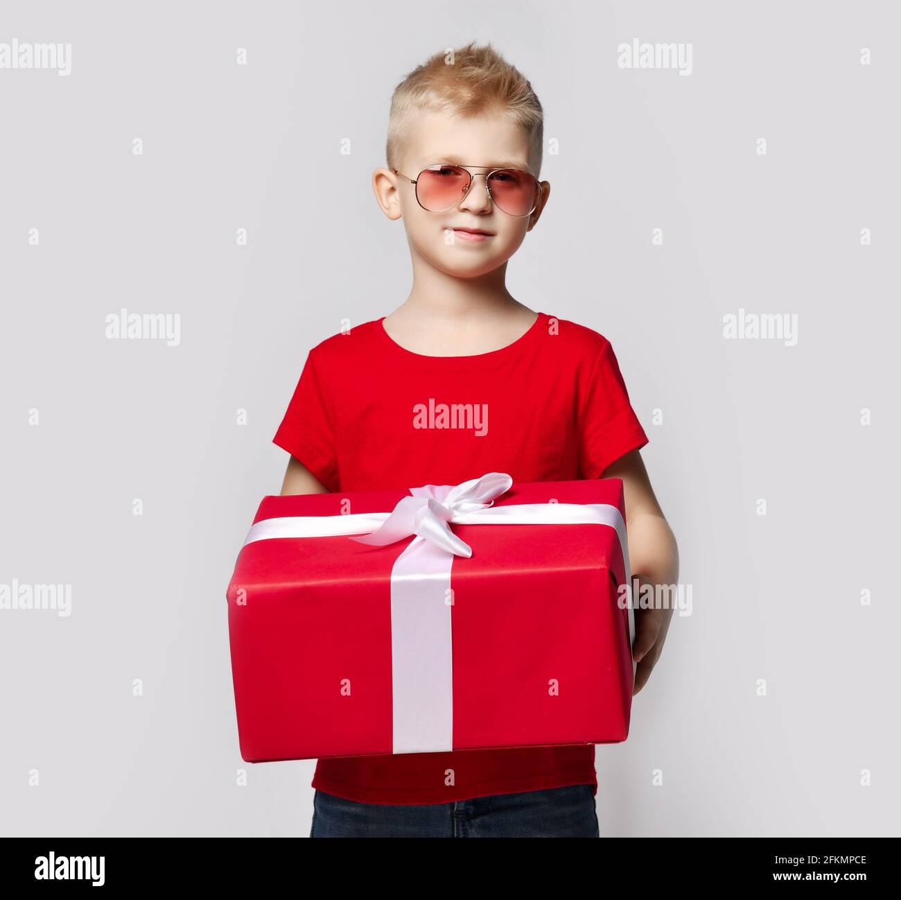 Stylish kid boy in red t-shirt and sunglasses stands holding big present for birthday gift box with ribbon in hands Stock Photo