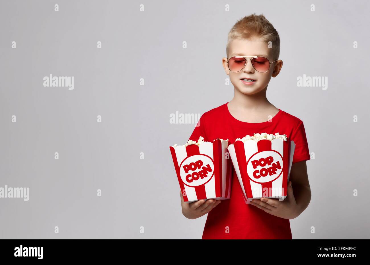 Stylish kid boy in red t-shirt and sunglasses stands holding two popcorn boxes in hands, ready for dating in cinema Stock Photo