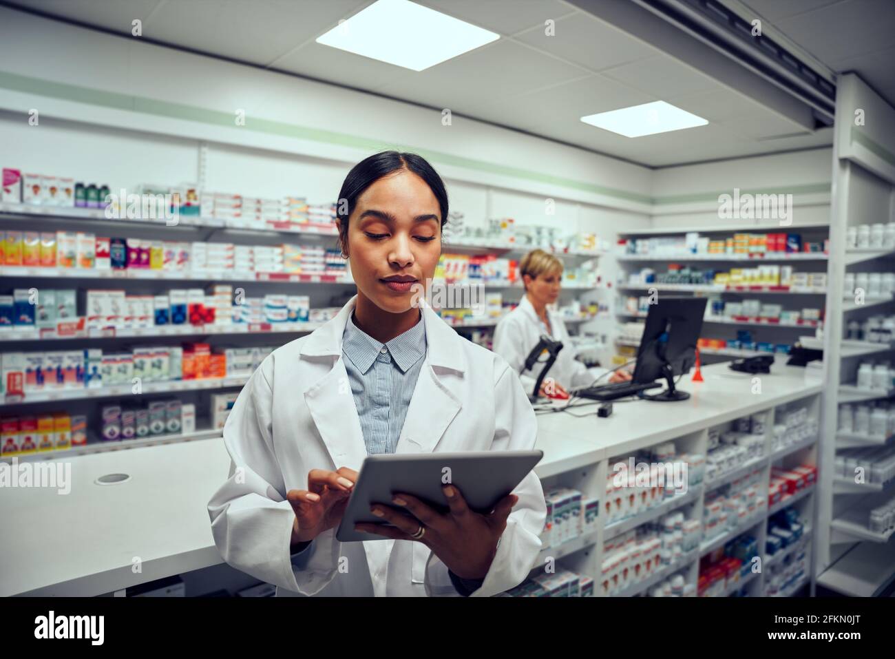 Portrait of young female working in pharmacy wearing labcoat standing while using digital tablet to check online order Stock Photo