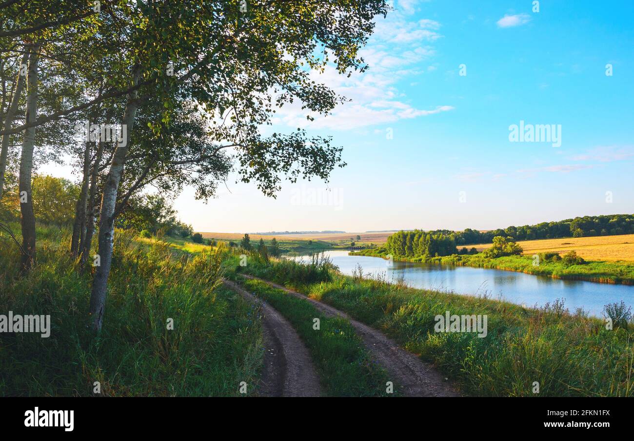 Summer serene landscape with calm river and rural dirt road at sunrise. Stock Photo