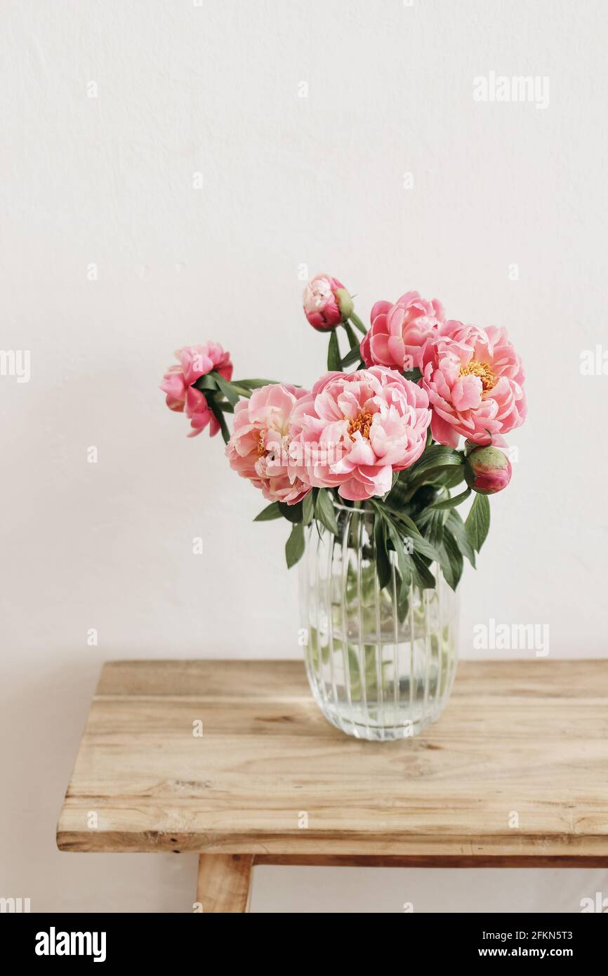 Floral still life scene. Pink peonies flowers, bouquet in glass vase on wooden table. White wall. Selective focus, blurred background. Wedding Stock Photo