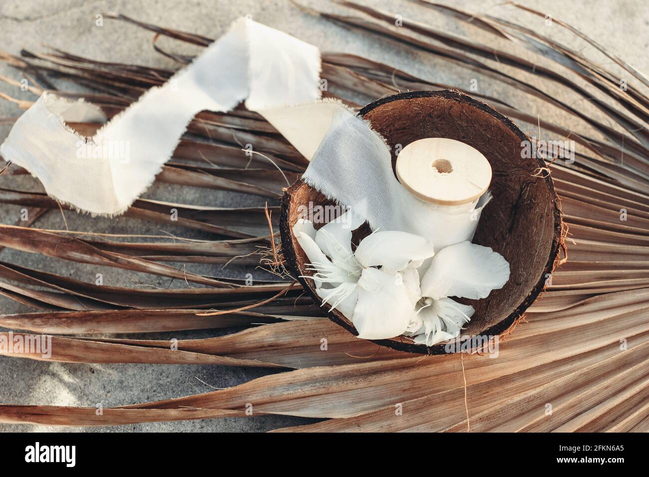 Summer tropical lifestyle scene. Oleander blossoms and white silk ribbons spools in coconut shell. Dry palm leaves. Grunge concrete background in Stock Photo