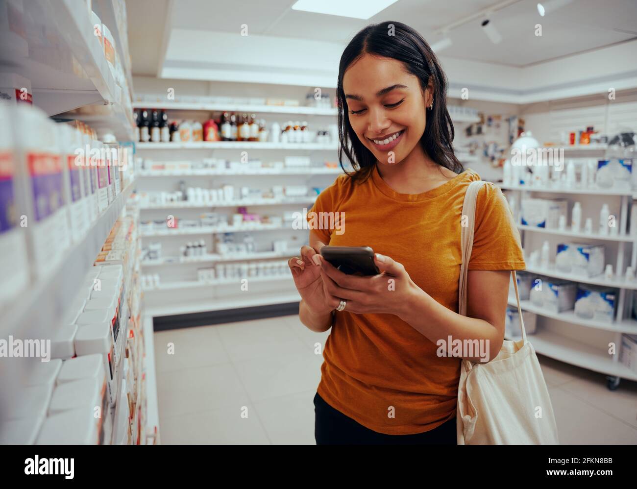 Young woman smiling while using smartphone standing against shelf in pharmacy Stock Photo