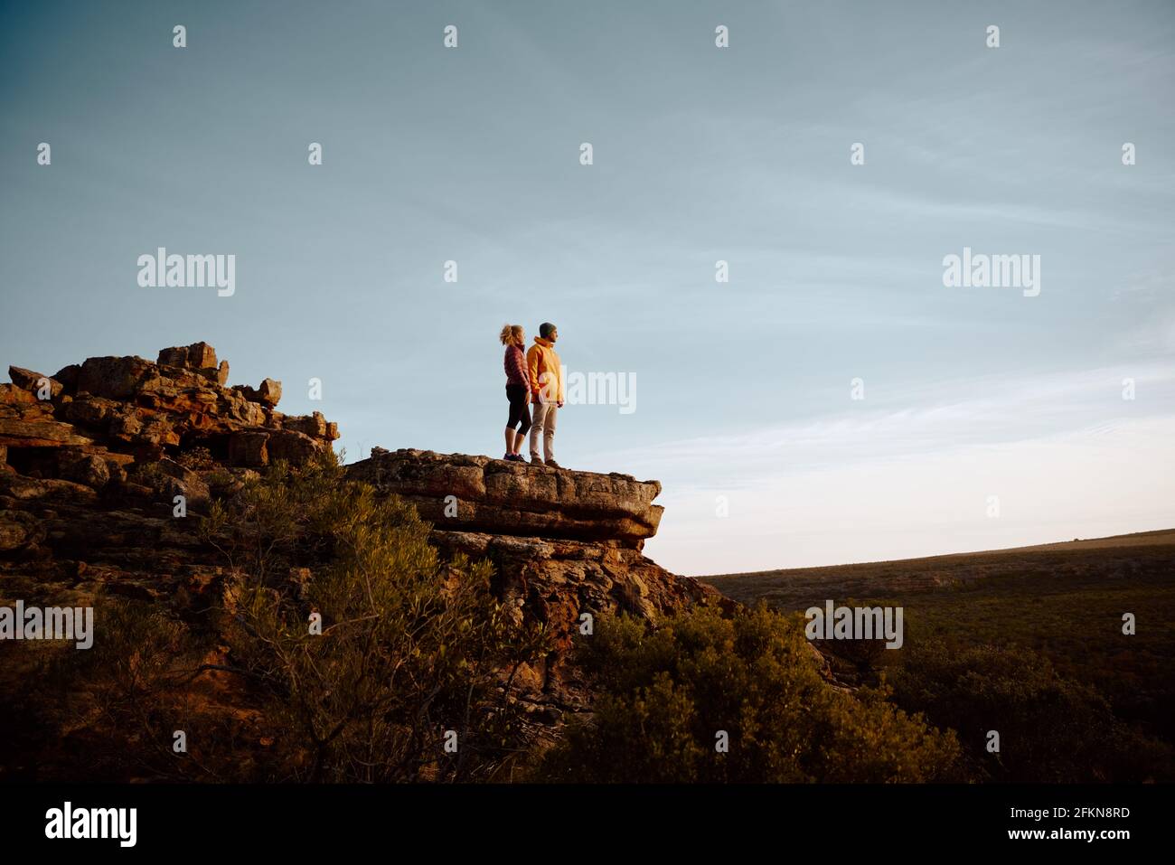 Low angle view of young man and woman standing looking at beautiful view after hiking standing at mountain cliff peak Stock Photo