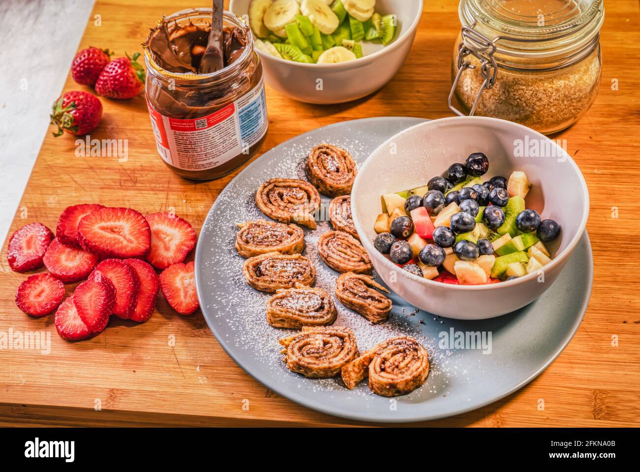 Chocolate hazelnut spread or Nutella cream crepe rolls with fresh fruits like strawberries, kiwi, banana and blueberries on rustic wooden board Stock Photo