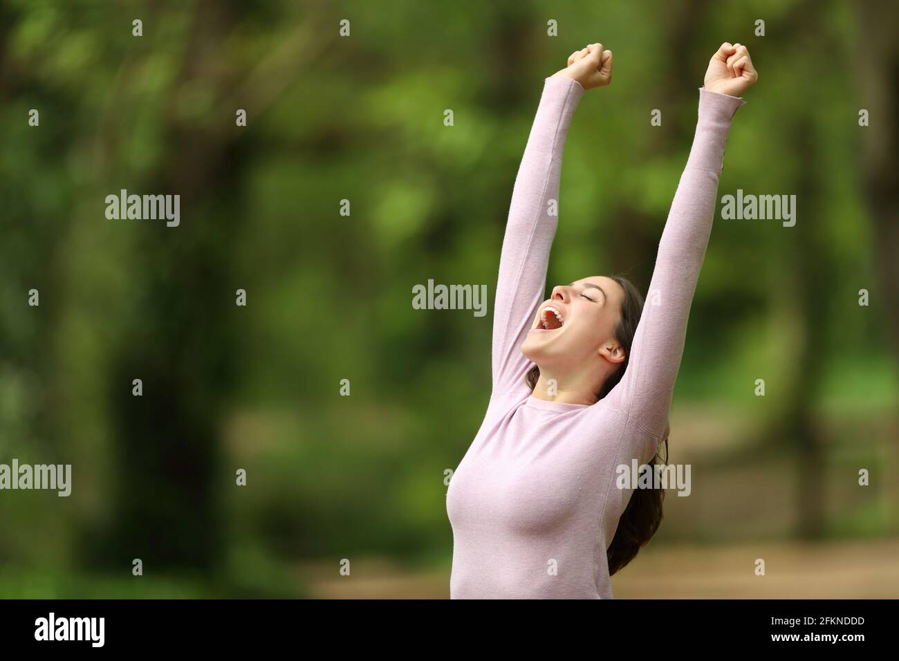 Excited woman raising arms and screaming celebrating in a green forest ...