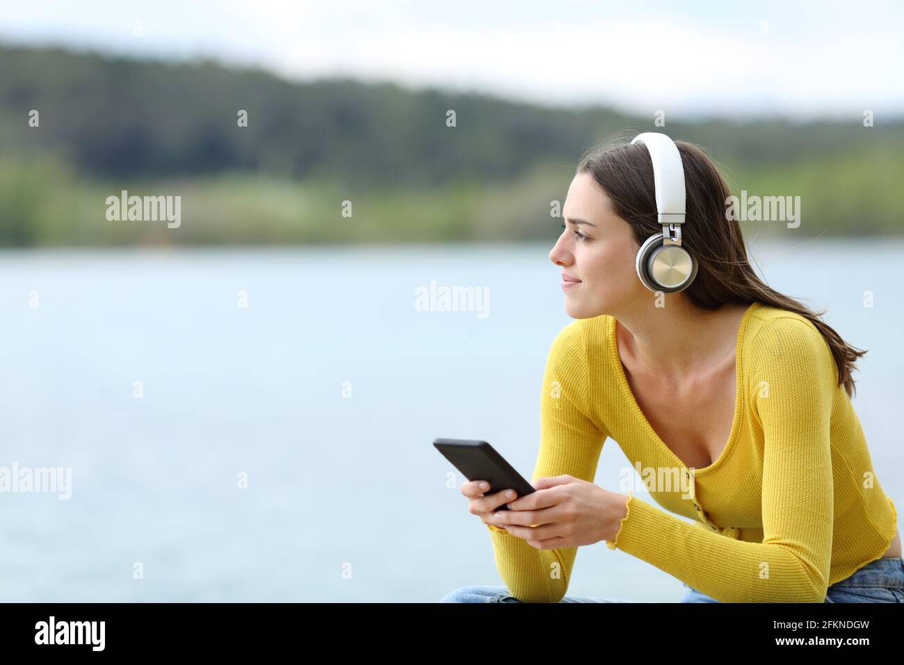 Satisfied woman listening to music with wireless headphones looking away in a lake Stock Photo