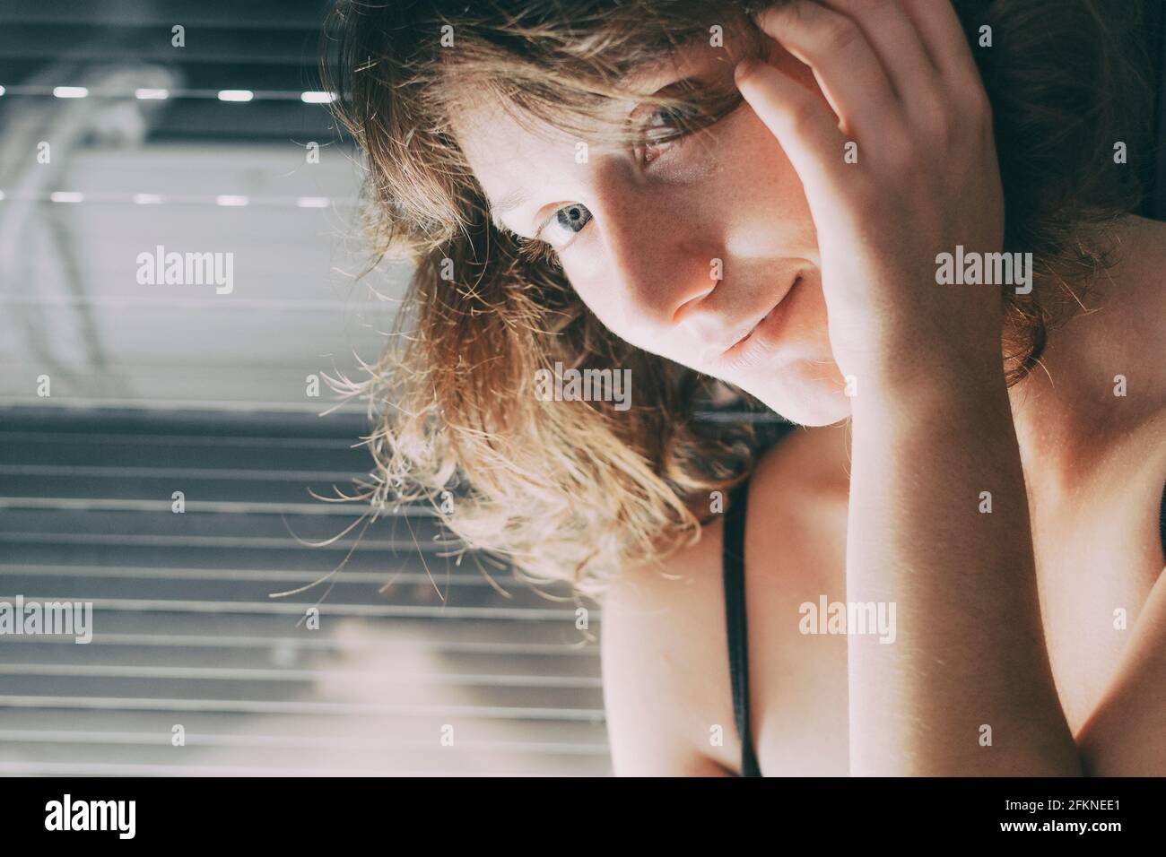 young woman with blue eyes and short hair inside her house, sitting next to the closed blind where her body is reflected in the window Stock Photo