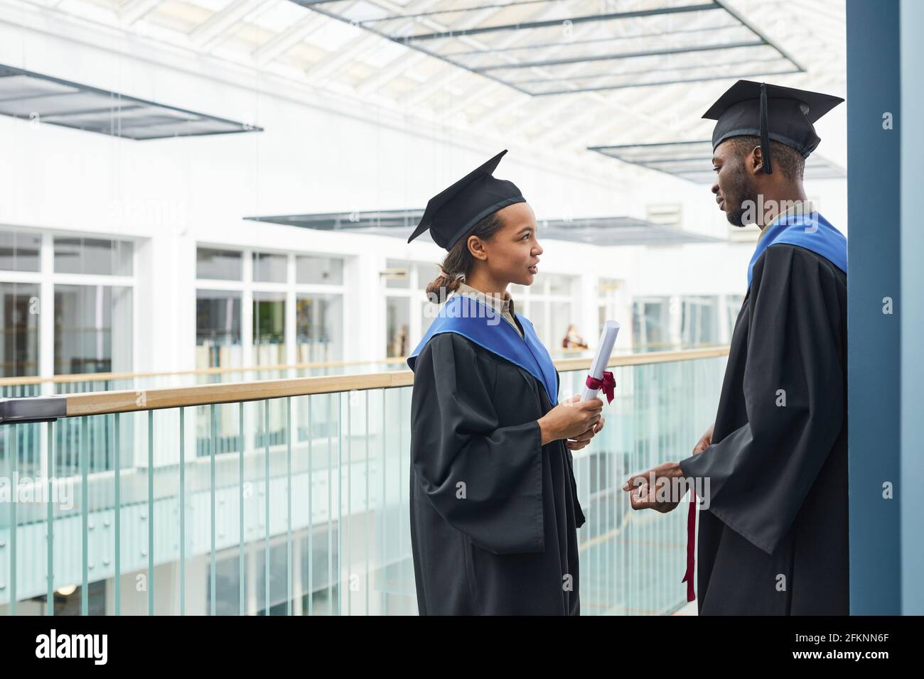 Side view at two young people wearing graduation gowns chatting indoors ...