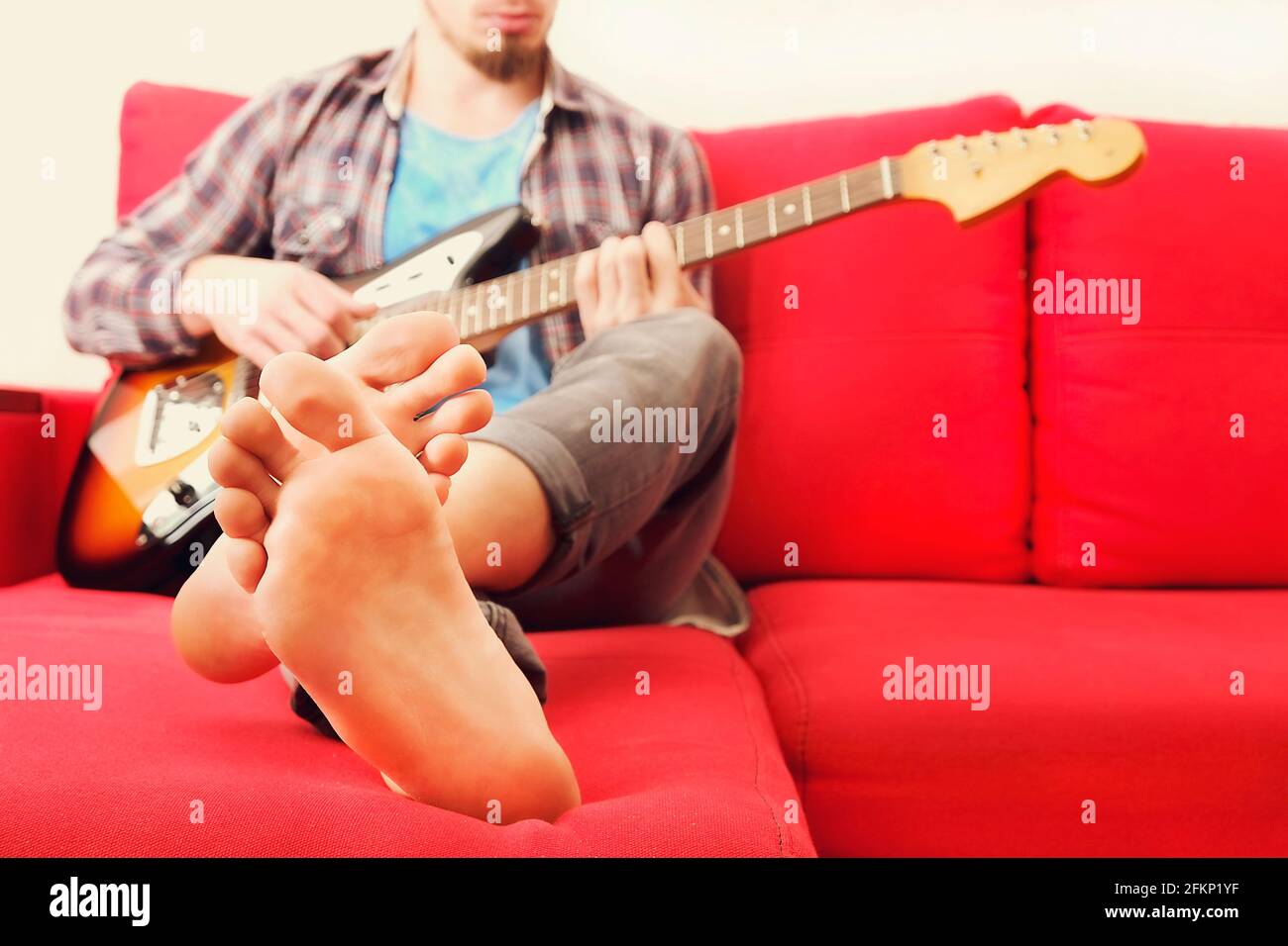 Barefoot man in checkered plaid shirt sitting on red sofa by the window playing offset electric guitar, mahogany neck. Rock guitarist plays jazz impro Stock Photo