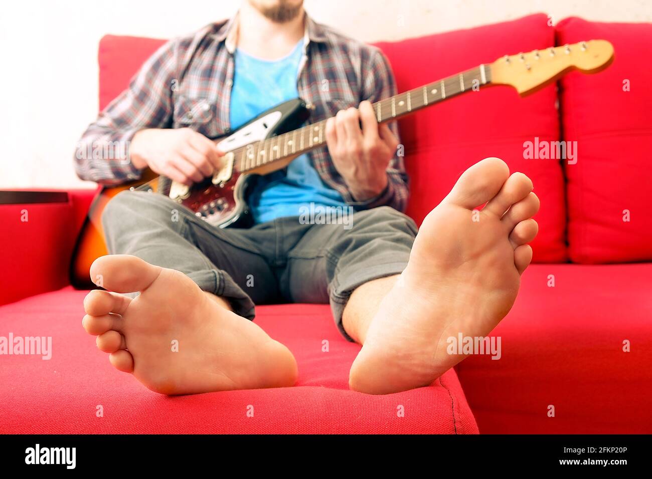 Barefoot man in checkered plaid shirt sitting on red sofa by the window playing offset electric guitar, mahogany neck. Rock guitarist plays jazz impro Stock Photo