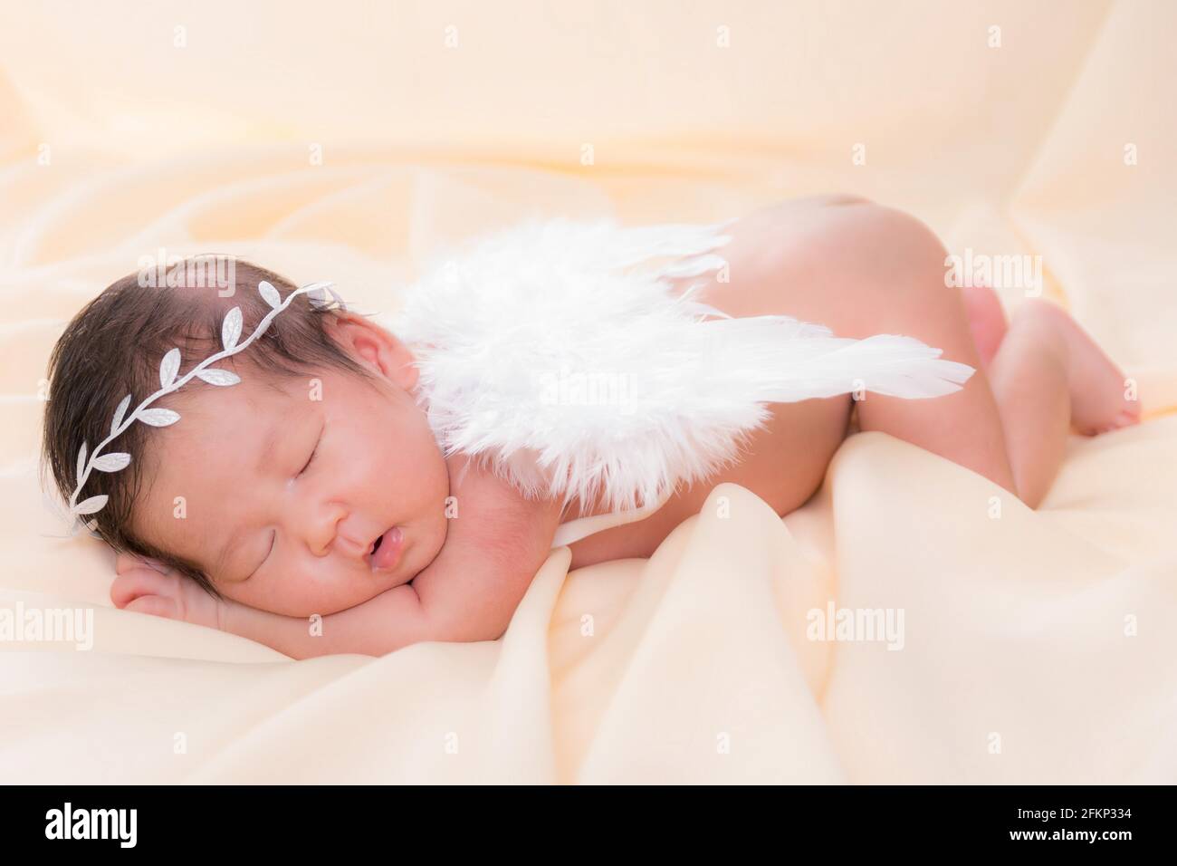 Portrait of a one month old sleeping, newborn baby girl. She is wearing a white crown headband, angel wings and sleeping on a cream blanket. Concept p Stock Photo