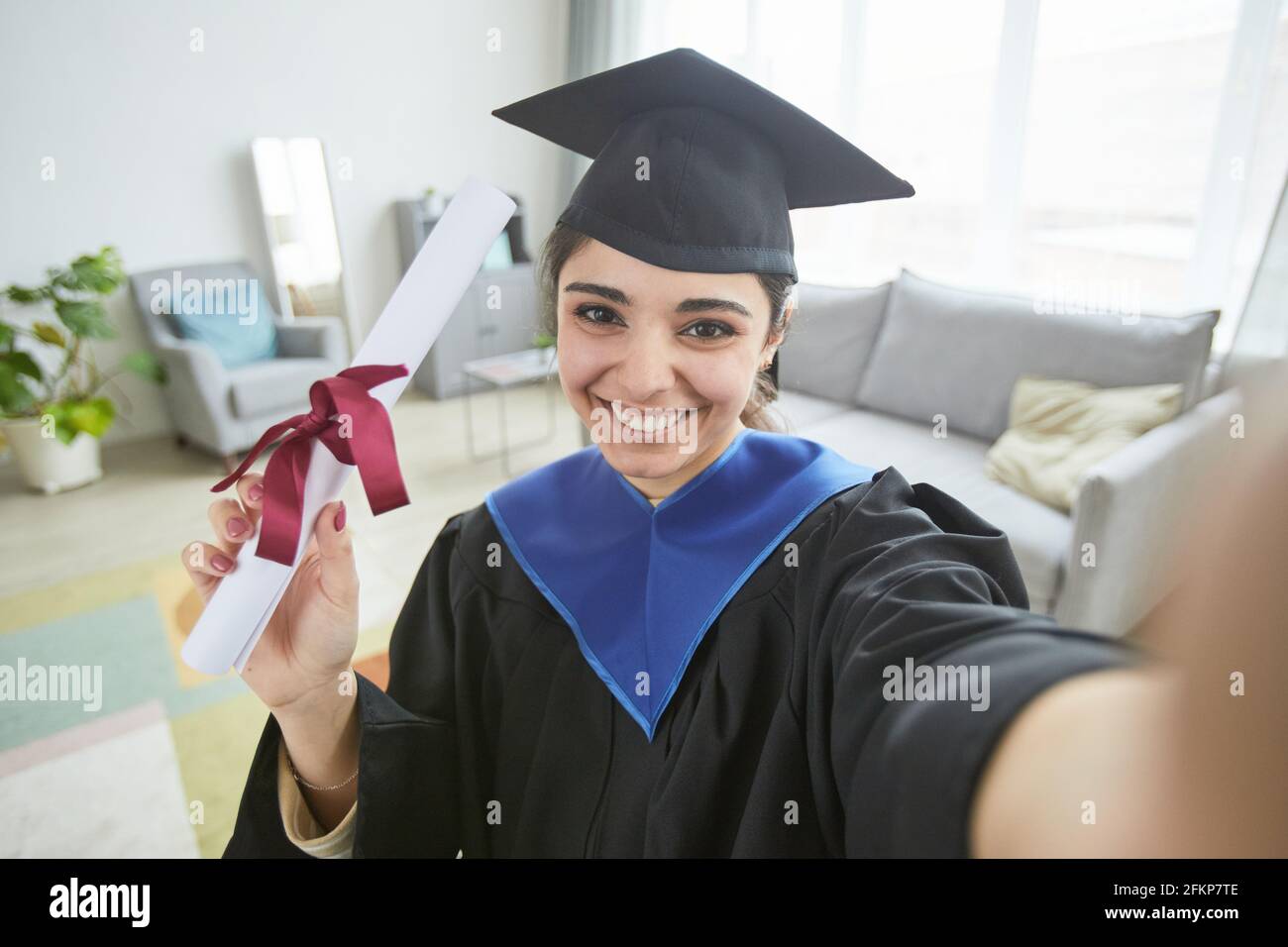 Smiling young woman wearing graduation gown while taking selfie at home or video blogging Stock Photo