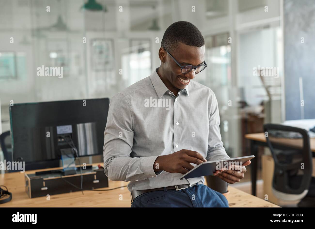 Smiling African businessman leaning on an office desk using a tablet ...