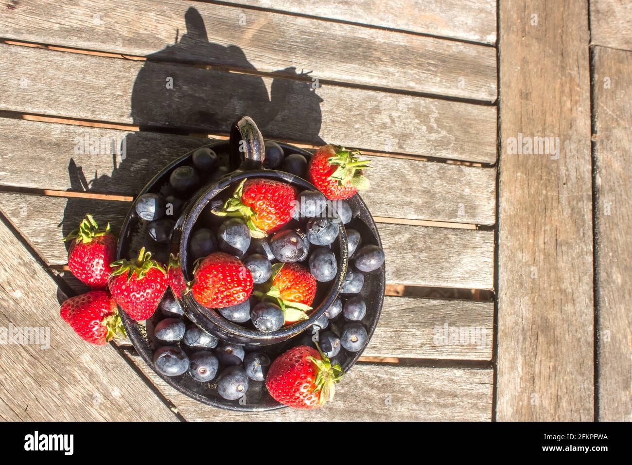 A lay-flat composition of a teacup filled to overflowing with strawberries and blueberries on a weathered, wooden surface Stock Photo