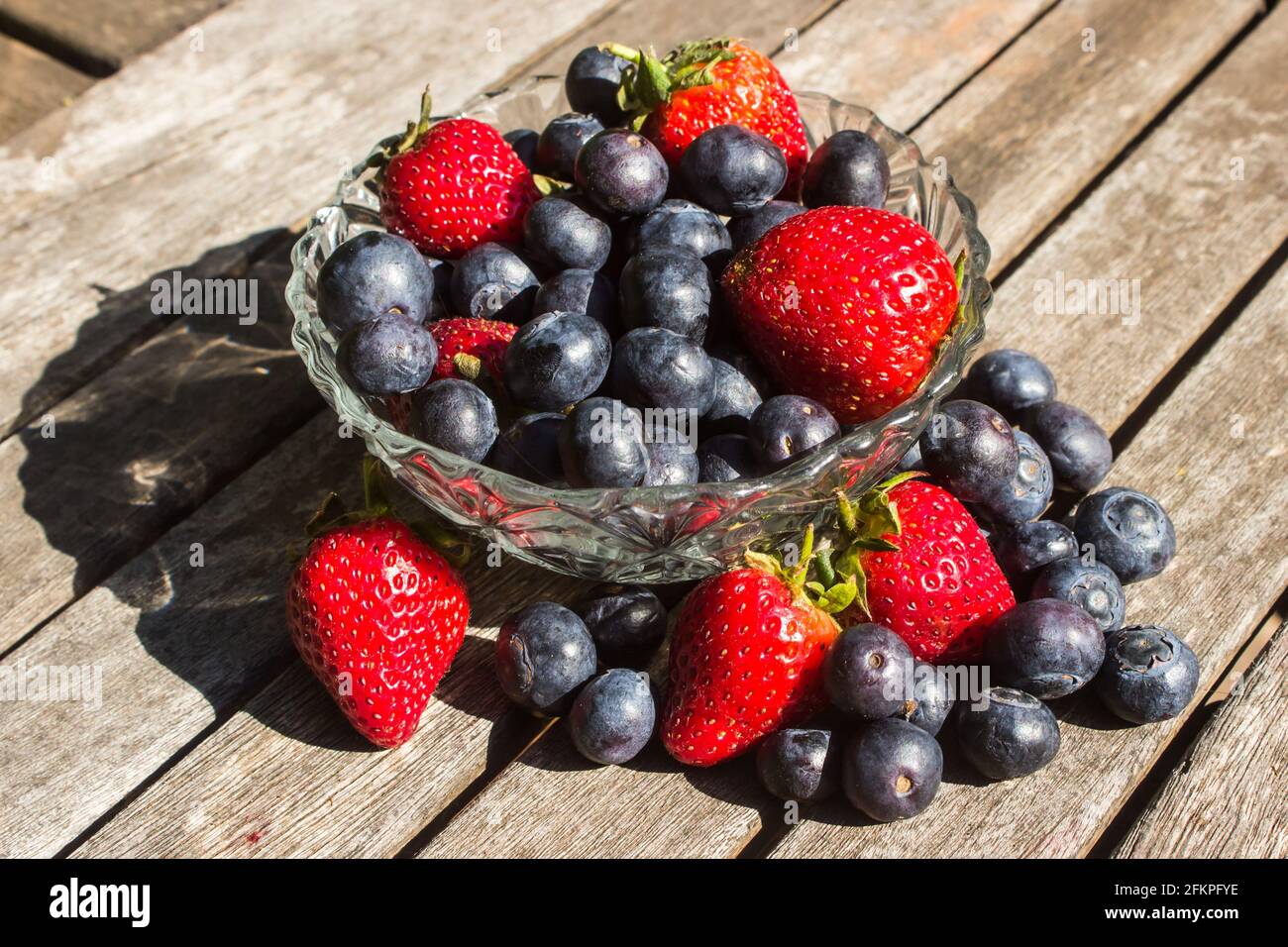 A glass bowl, filled to overflowing with bright red strawberries and indigo-colored blueberries on a weathered wooden surface Stock Photo